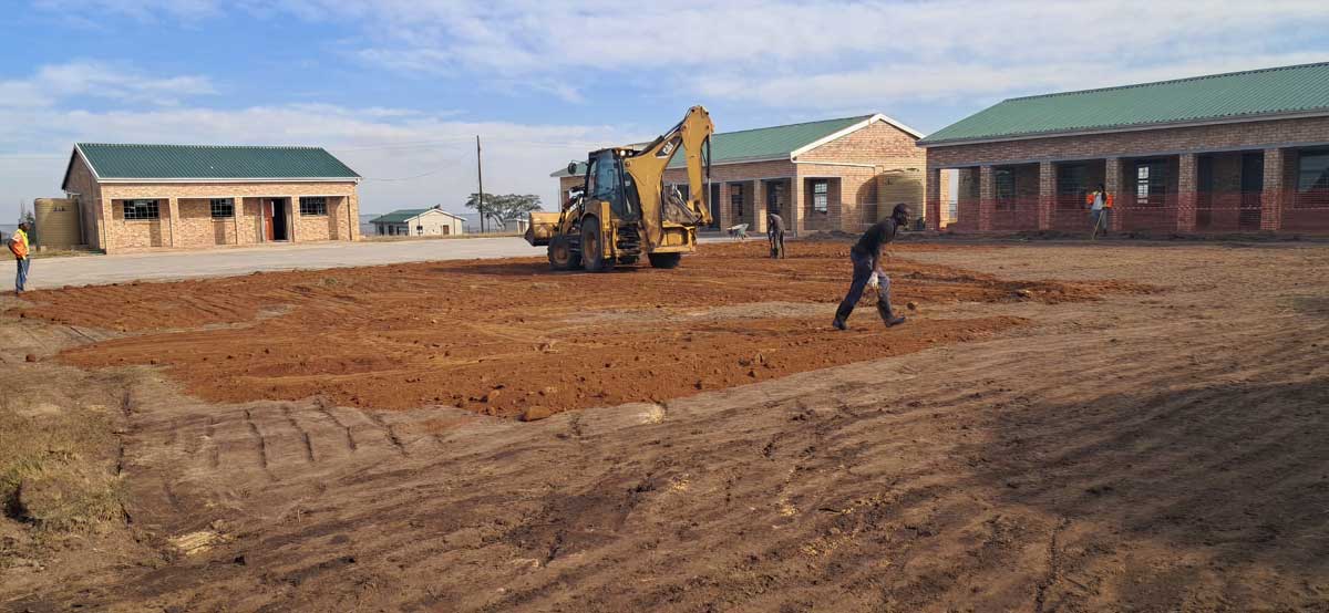 about-us-image Construction workers leveling the ground with a backhoe loader near newly built brick buildings.